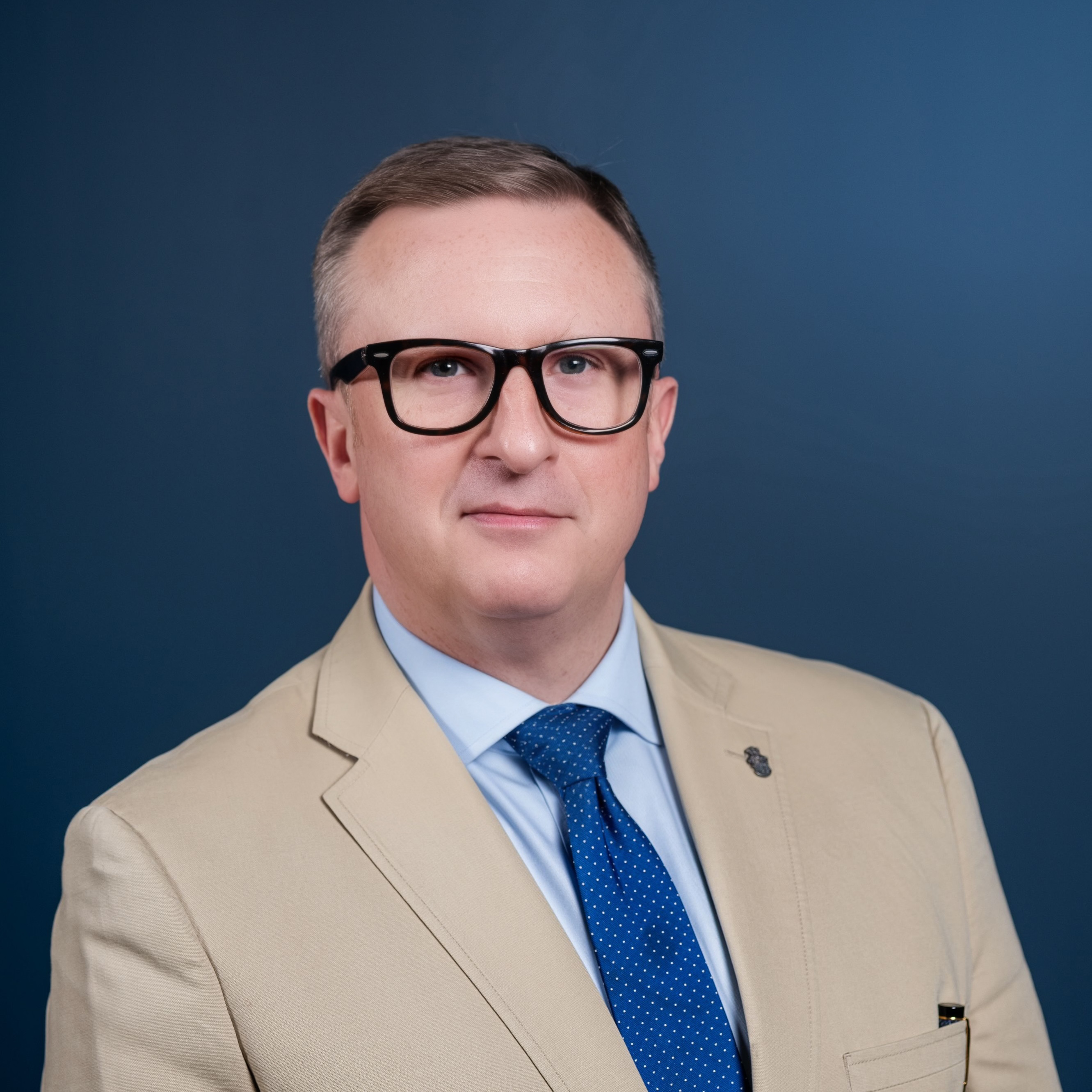 Man in tan blazer and blue tie in front of dark blue background